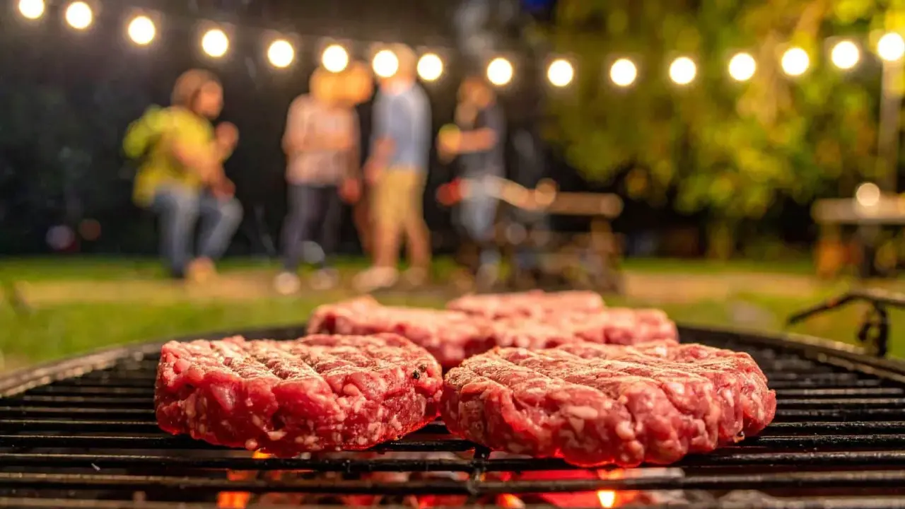 Frische Burger-Patties liegen auf dem Grill. Im Hintergrund feiern Freunde unter Lichterketten. So macht man Hackfleisch selber für den perfekten Grillabend.