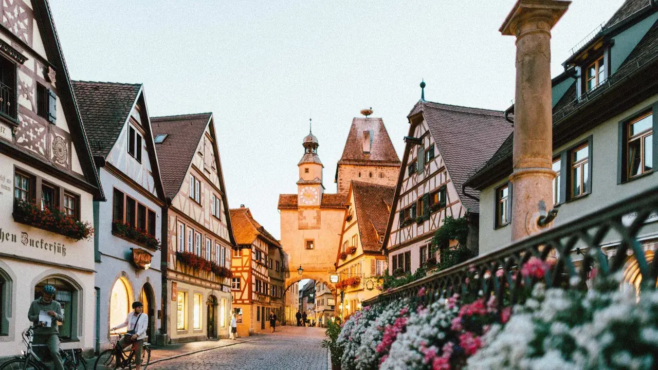 A charming street in gay Germany, with historic buildings, cobblestones, and flowers. Two people on bikes pause to take in the view.