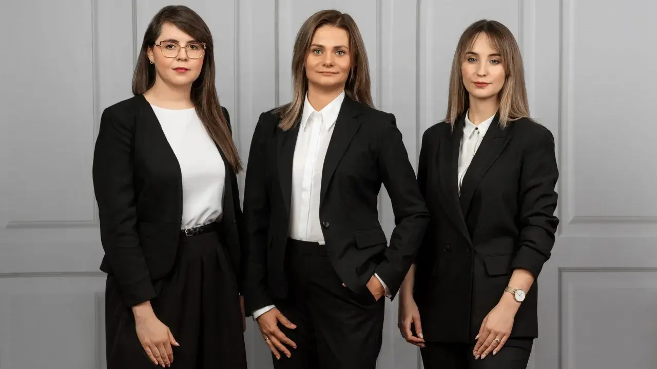 Trzy kobiety w czarnych garniturach stoją przed szarą ścianą.