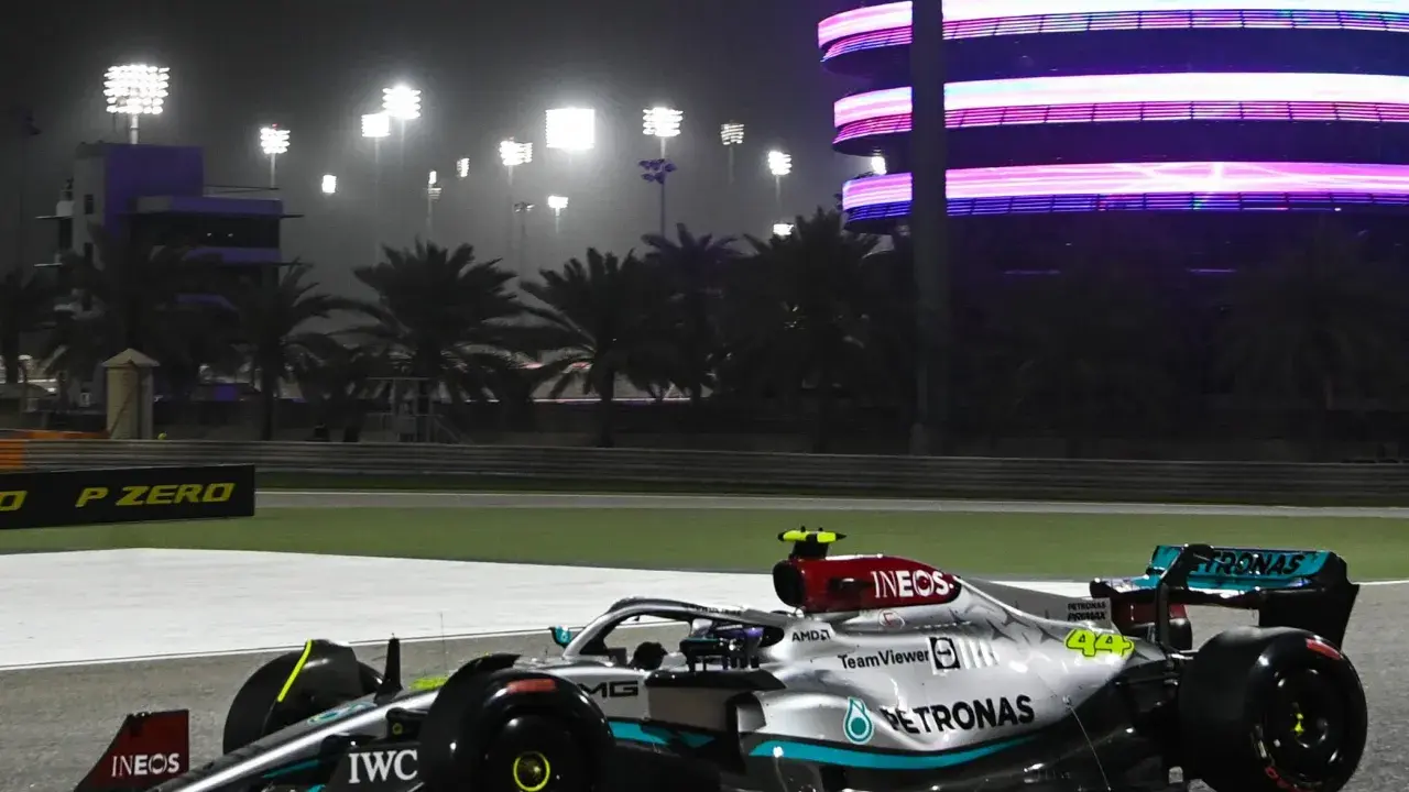 Silberner F1-Rennwagen mit Petronas-Sponsoren fährt bei Nacht auf der Rennstrecke in Bahrain. Flutlicht und ein beleuchtetes Gebäude im Hintergrund.