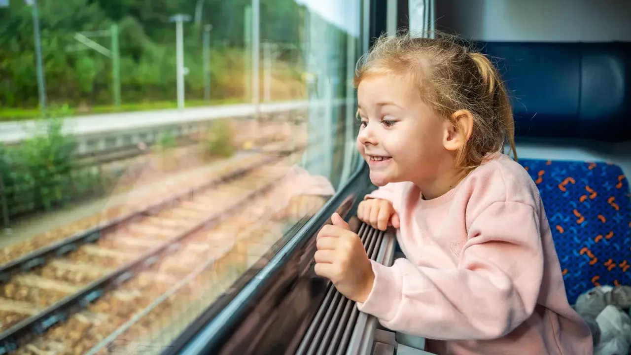 Uśmiechnięta dziewczynka patrzy przez okno pociągu. Zastanawia się, od ilu lat można jeździć samemu pociągiem, podziwiając mijane krajobrazy.