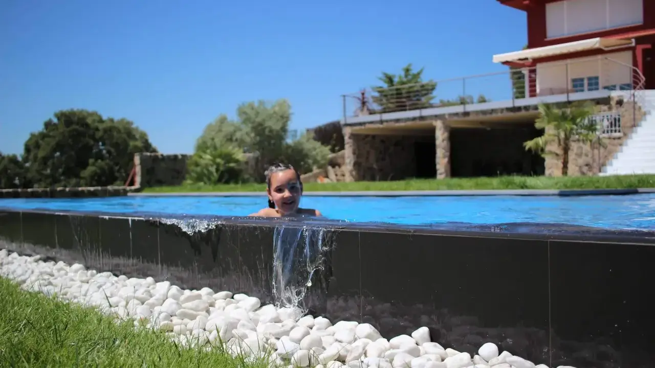 Niña sonriente en una piscina desbordante, con agua cayendo sobre piedras blancas y césped verde. Al fondo, una casa moderna y árboles frondosos bajo un cielo azul.