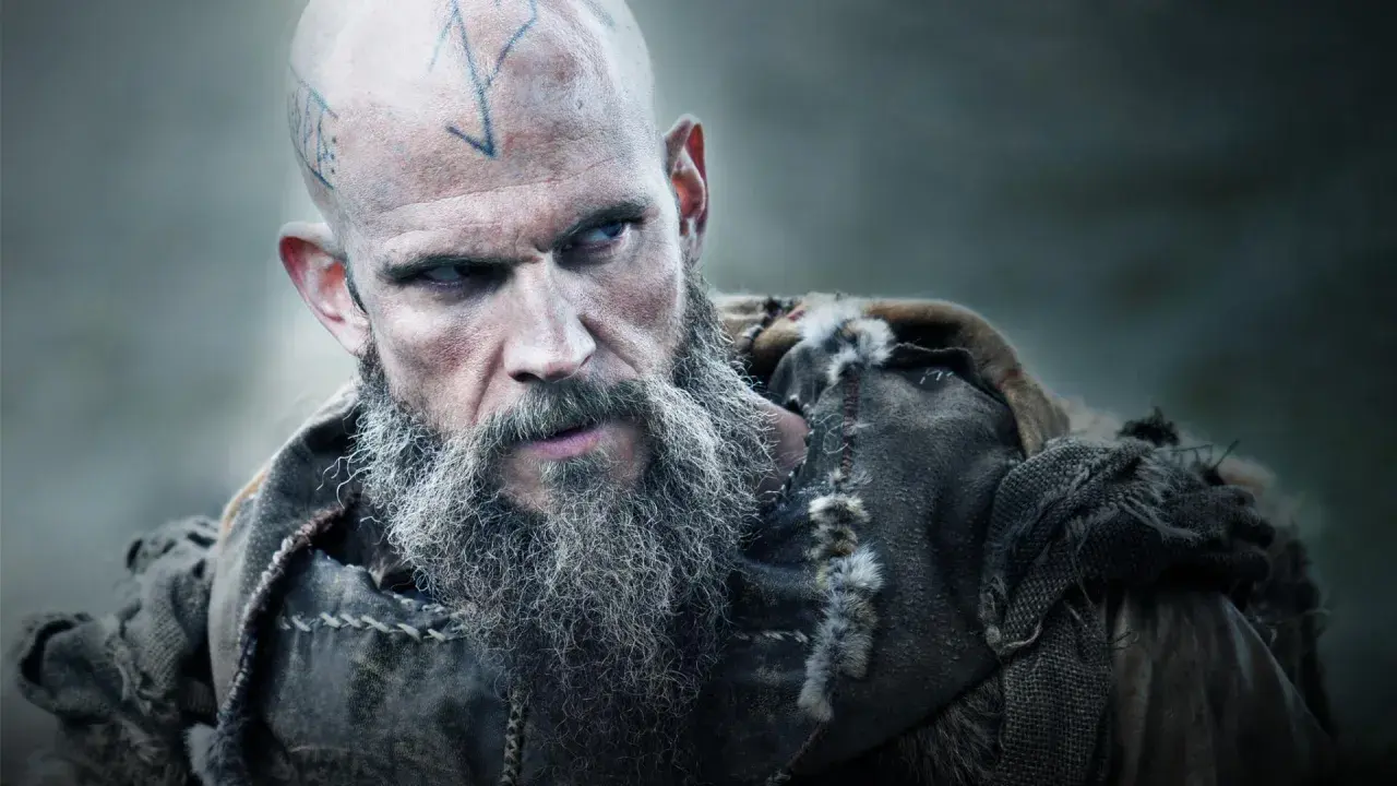 Uhtred z Bebbanburga, wojownik z tatuażami na głowie i długą, siwą brodą, spogląda groźnie.