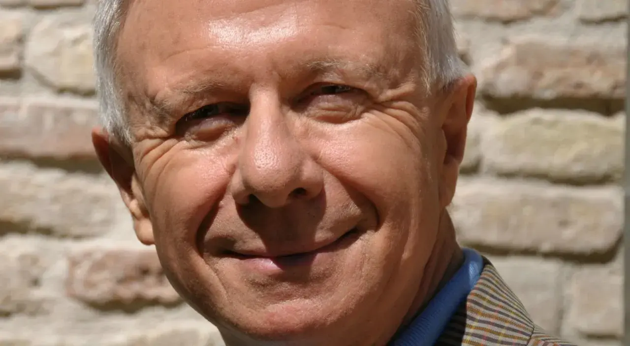 Primo piano sorridente di padre Alberto Maggi, con rughe d'espressione e capelli grigi, sullo sfondo di un muro di mattoni.
