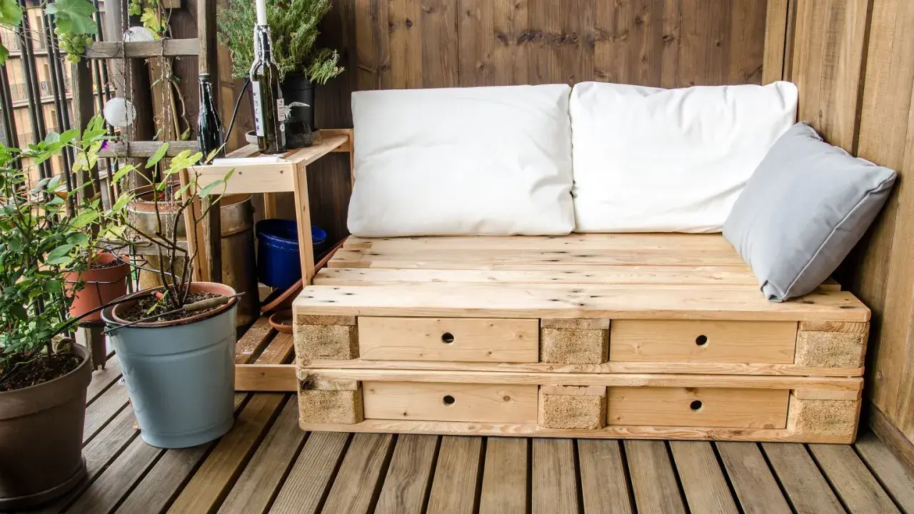 Meble z palet na balkon: wygodna sofa z szufladami, białe i szare poduszki, rośliny w doniczkach, drewniana ściana i podłoga.