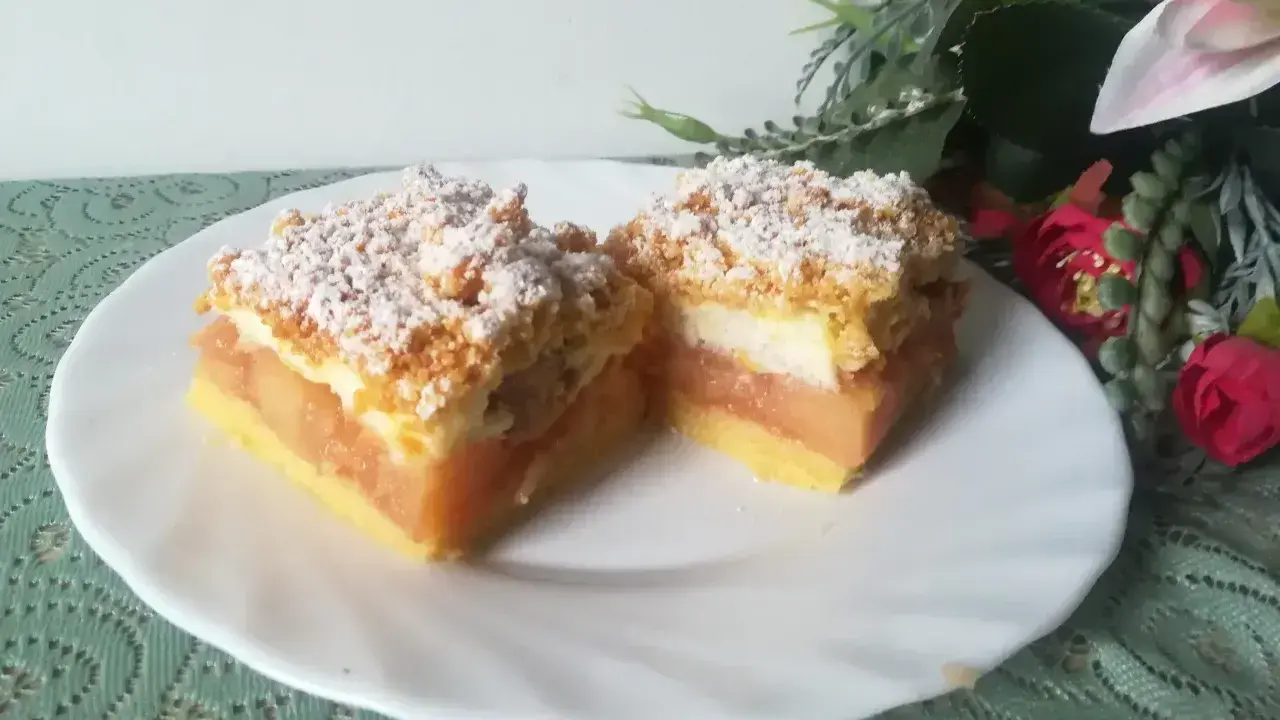 Pyszna szarlotka z bezą i kruszonką, z jabłkami i cukrem pudrem, na białym talerzu, obok kwiatów.