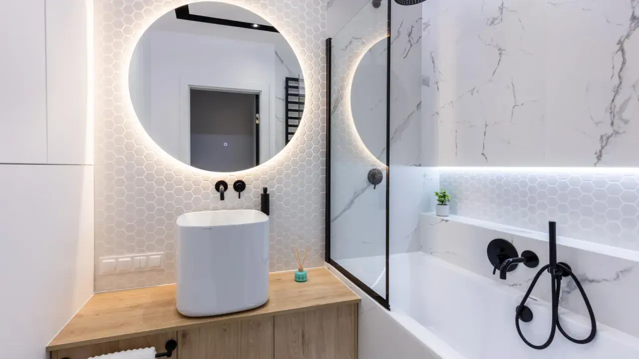 Elegante bagno senza finestra con lavabo bianco, specchio rotondo illuminato e doccia con finiture nere.