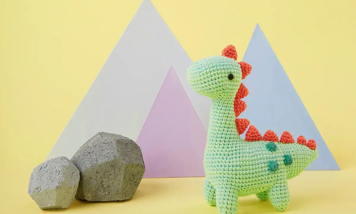 Adorable dinosaurio crochet verde con púas naranjas y detalles verdes. Dos rocas grises y formas geométricas de colores pastel al fondo.