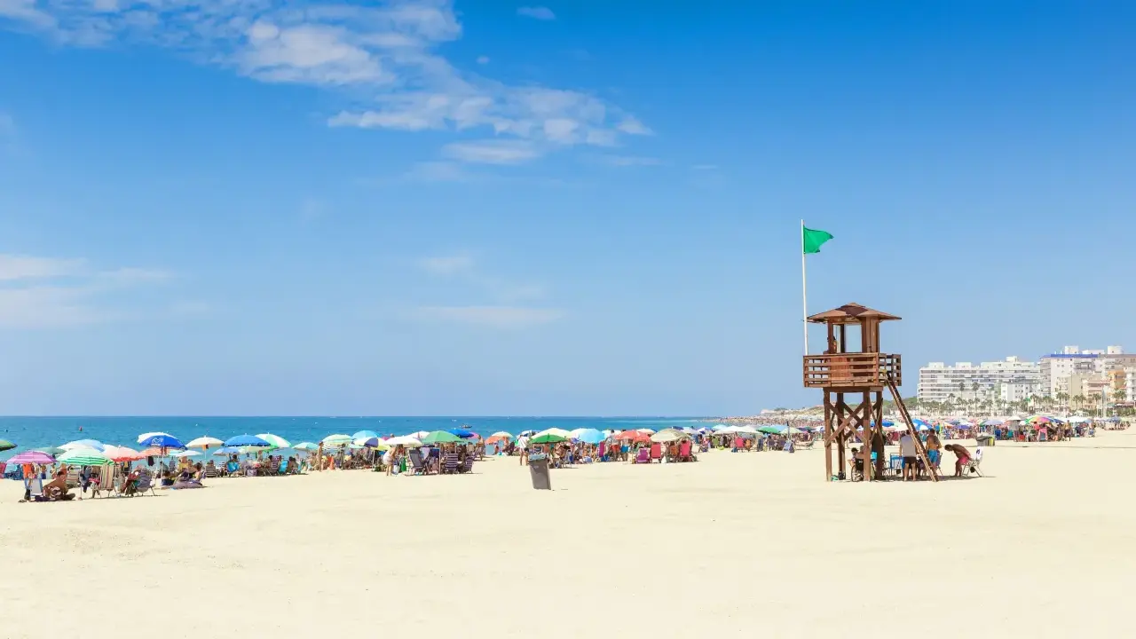 Sonniger Strand in Rota, Spanien, mit vielen bunten Sonnenschirmen und Menschen. Ein Rettungsschwimmerturm mit grüner Flagge steht am Meer.
