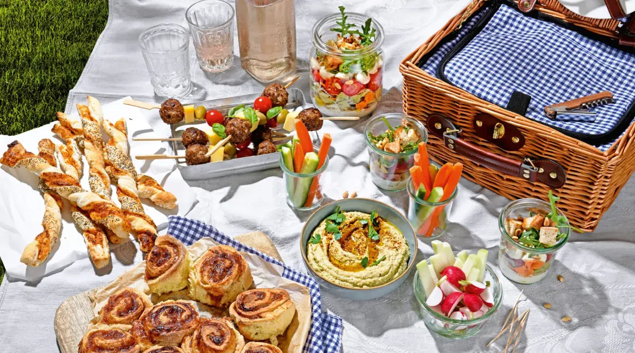 Leckere Picknick-Snacks: Fleischbällchen-Spieße, Gebäckstangen, Hummus, Gemüsesticks und süße Schnecken auf einer Decke.