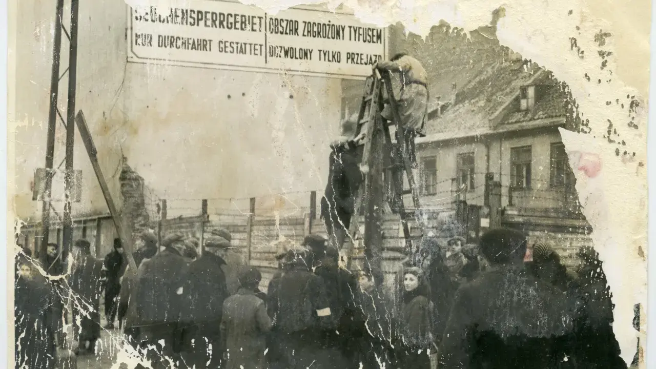 1941. Ludzie stoją przed znakiem "Obszar zagrożony tyfusem". W tle budynki i druty kolczaste.