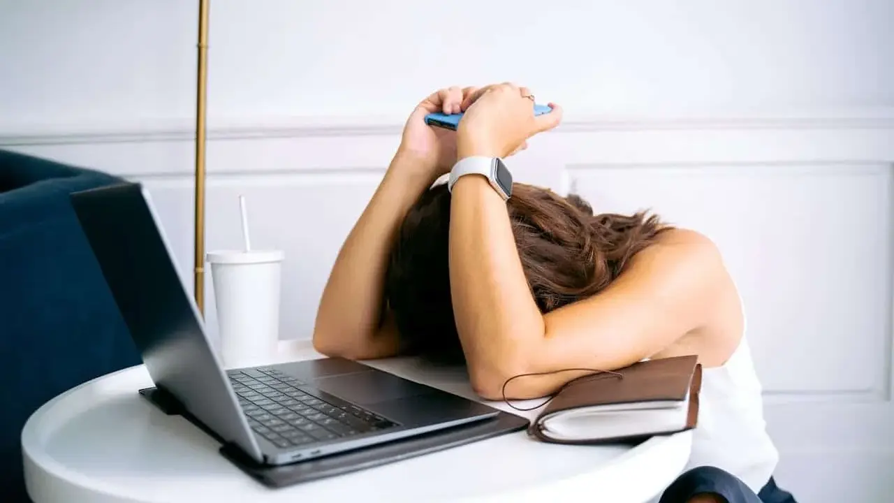 Personne épuisée, tête sur les bras, téléphone à la main, devant un ordinateur portable. L'effet écran de la fatigue est palpable.