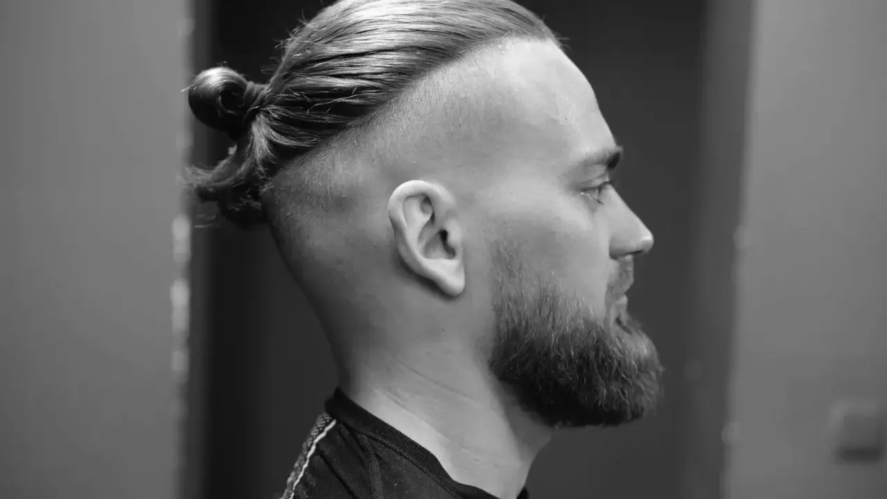 Mężczyzna z brodą i fryzurą na samuraja, z włosami upiętymi w kok na czubku głowy.