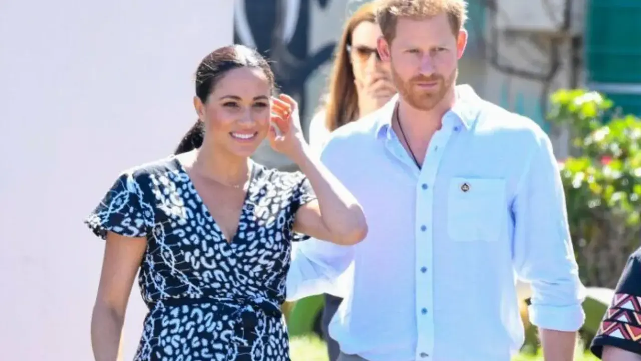 Meghan och Harry ler glatt under ett besök utomhus. Meghan bär en mönstrad klänning och Harry en ljusblå skjorta.