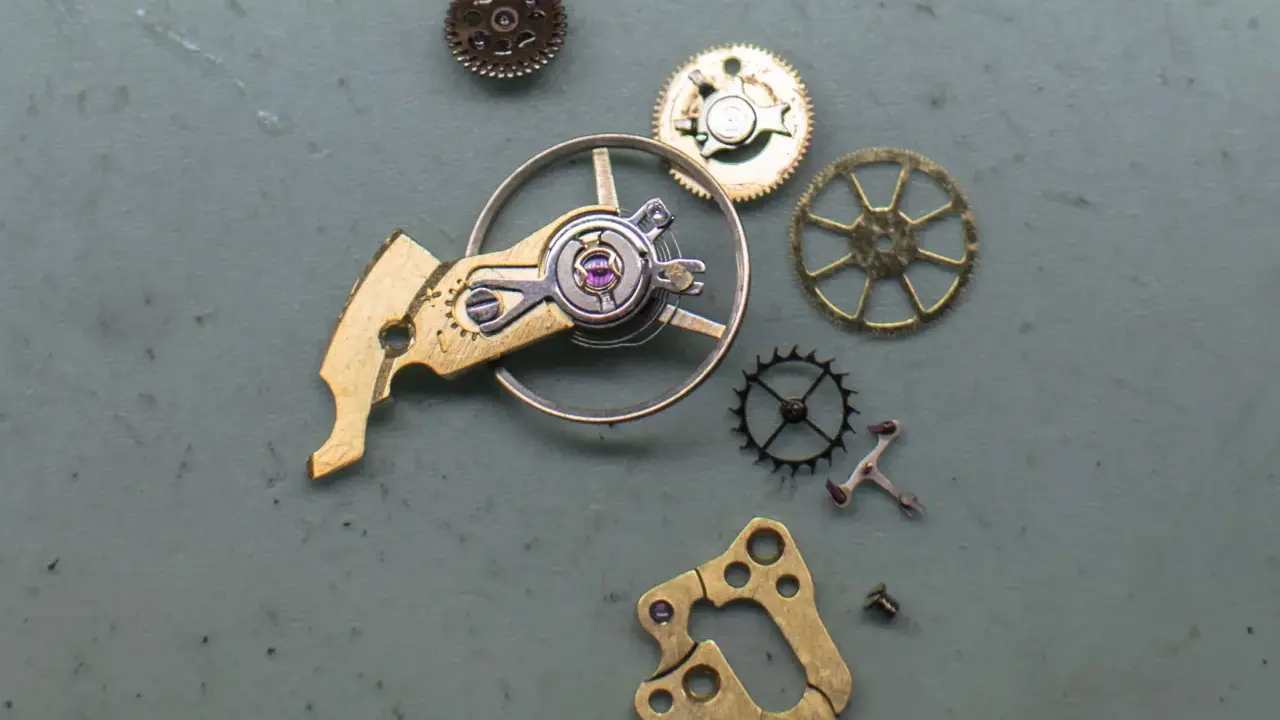 Parti di un orologio: ingranaggi, un bilanciere e altri componenti metallici disposti su una superficie grigia.