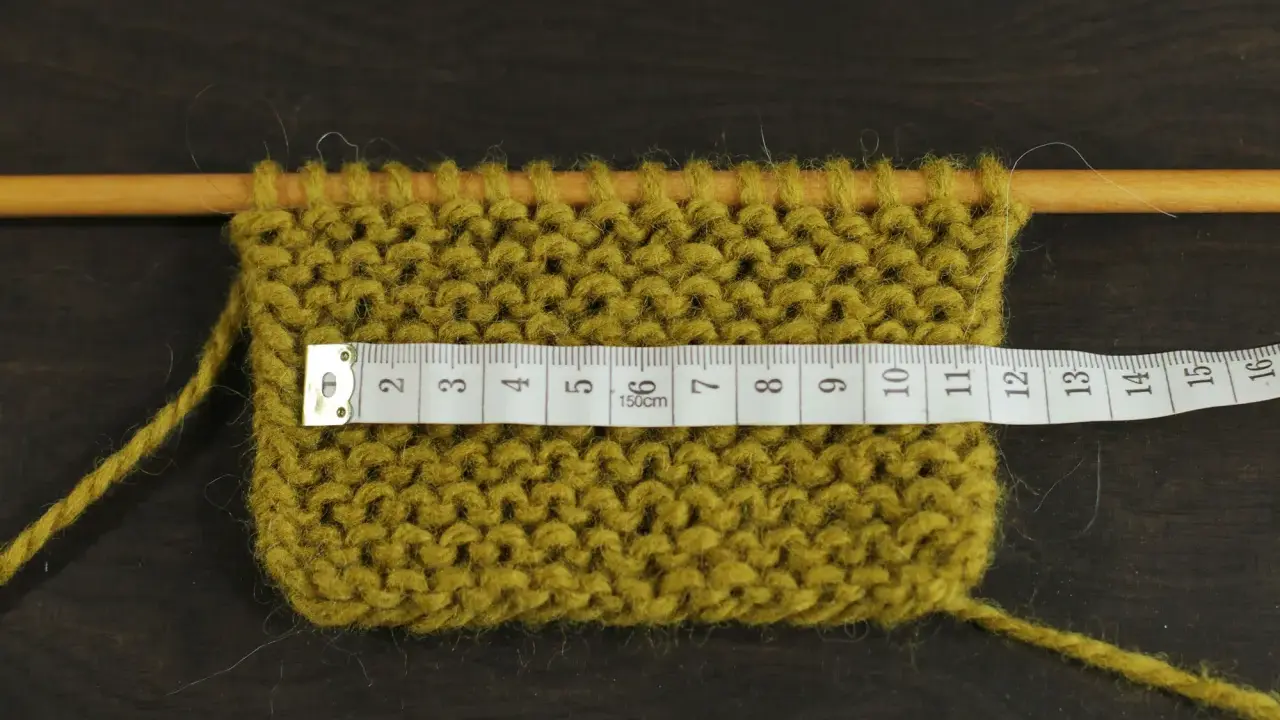 Knitted swatch with a measuring tape across it, showing stitches on wooden needles.