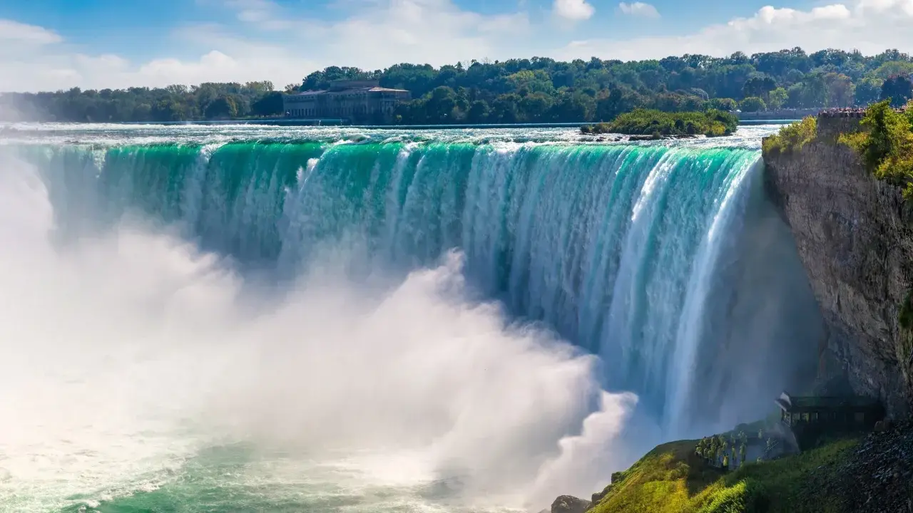 Potężny wodospad Niagara w całej okazałości. Woda spada kaskadami, tworząc mgłę. W tle drzewa i budynek.