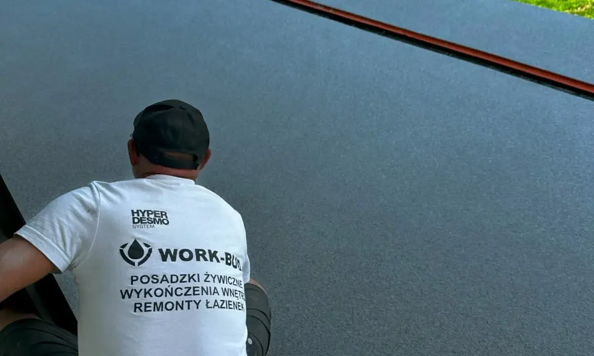 Pracownik w koszulce z napisem "POSADZKI ŻYWICZNE" wykonuje prace na ciemnej powierzchni.