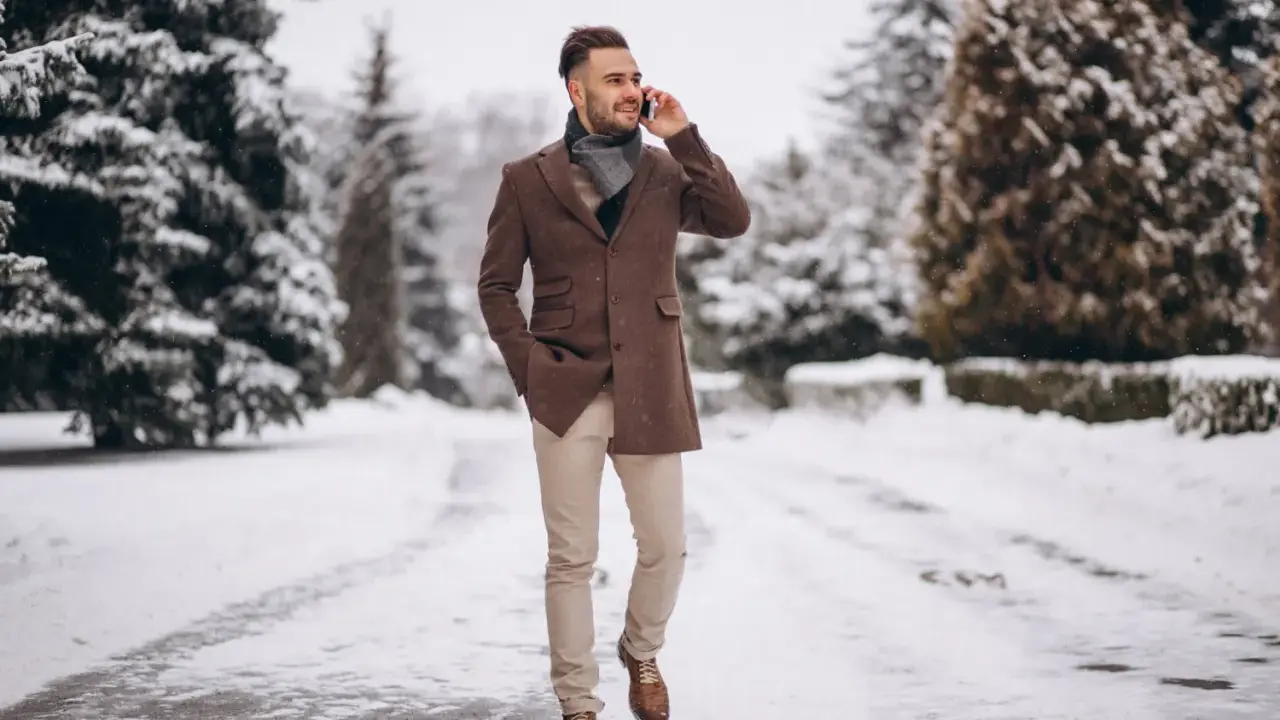 Man in stylish winter cocktail attire for men, wearing a brown coat and scarf, walks through a snowy park while on the phone.