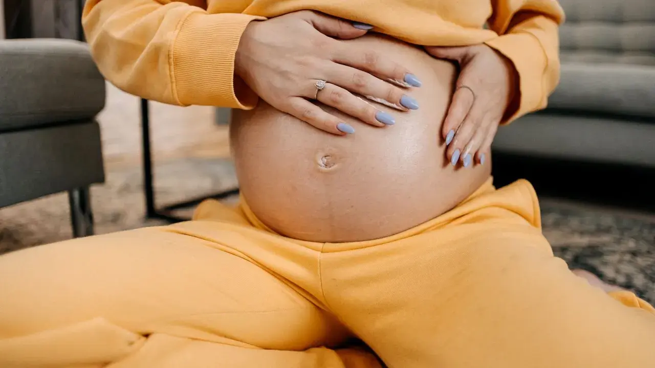 Schwangere Frau hält ihren Bauch, spürt den Druck im Unterleib. Gelber Jogginganzug, blaue Nägel.