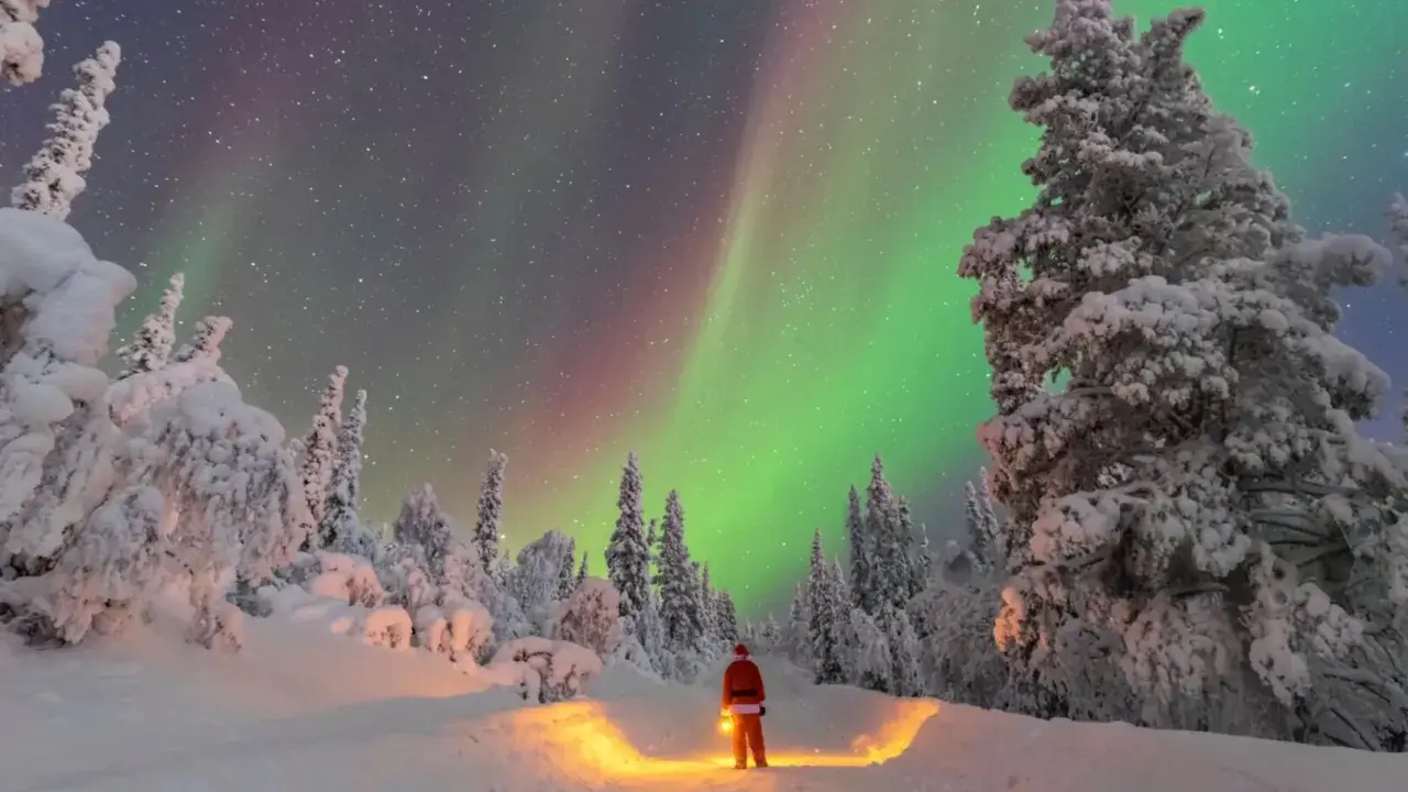 Un uomo con lanterna, vestito da Babbo Natale, ammira l'aurora boreale in Lapponia. Un paesaggio innevato e magico, perfetto per chi cerca dove andare in Lapponia.