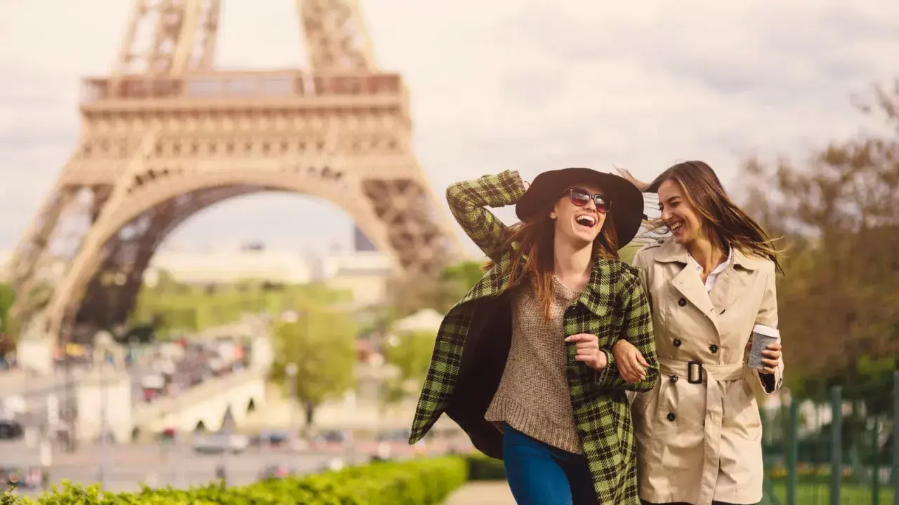 Due amiche ridono passeggiando a Parigi, con la Torre Eiffel sullo sfondo. Perfetto per capire cosa mettere in valigia per Parigi: cappotti leggeri, cappello e occhiali da sole.