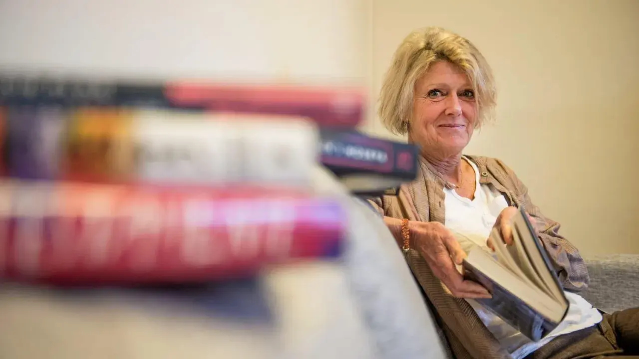 En kvinna med blont hår sitter med en bok i handen, omgiven av fler böcker. Hon ler mot kameran, som om hon delar en hemlighet från de fenomenala fruntimren på Grand Hotel.