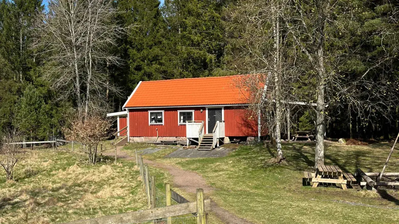 Rött torp med orange tak, omgivet av skog. En stig leder fram till torpet Freden, där picknickbord står redo för besökare.