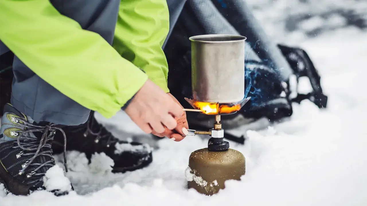 Tänd ett bensinkök i snön för att värma upp en kastrull. En person i en ljusgrön jacka och vandringskängor förbereder mat utomhus.