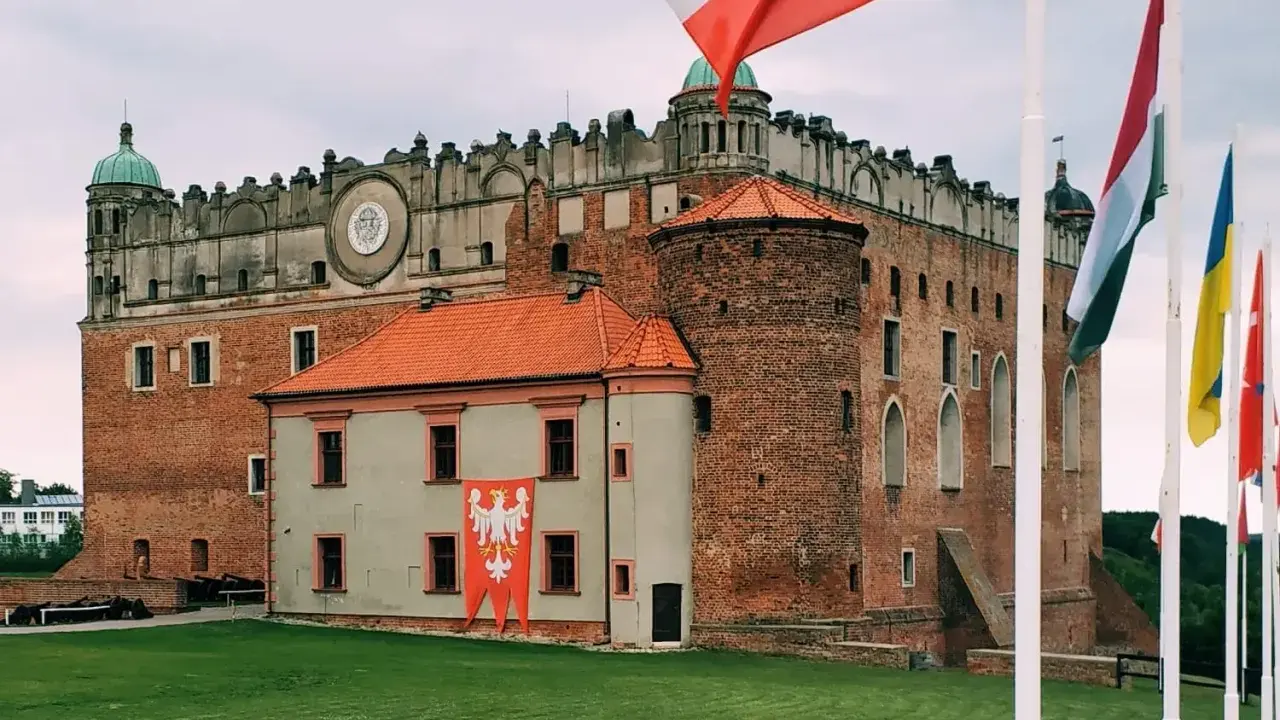 Majestatyczny zamek golubski z polską flagą i innymi flagami narodowymi na trawiastym dziedzińcu.