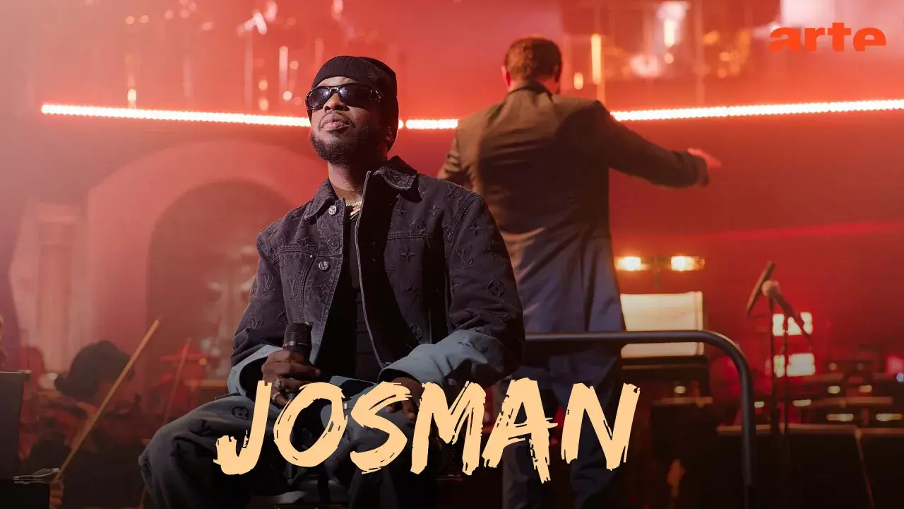 Josman, francuski rap artysta, siedzi na scenie z mikrofonem, w tle orkiestra i dyrygent.