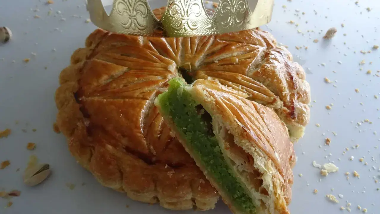 Galette des rois pistache dorée, une part coupée révèle sa garniture verte. Une couronne dorée trône sur ce délice.