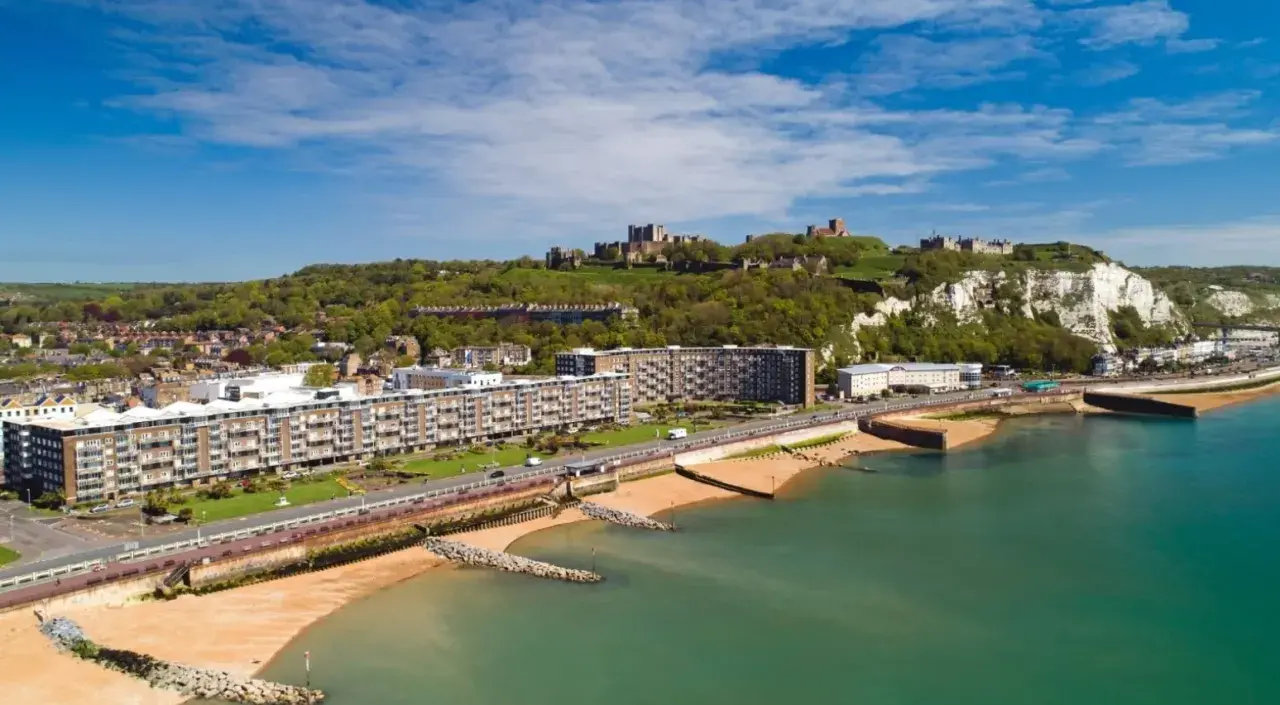 Vista aerea del porto di Dover con il castello in cima alla scogliera bianca. Edifici moderni e spiagge sabbiose si affacciano sul mare turchese.