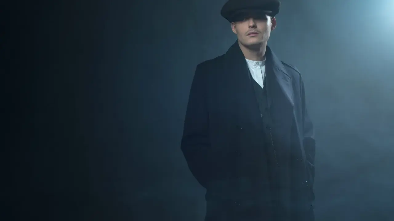 Młody mężczyzna w czapce i płaszczu, z charakterystyczną fryzurą w stylu Peaky Blinders, stoi w zadymionym mroku.