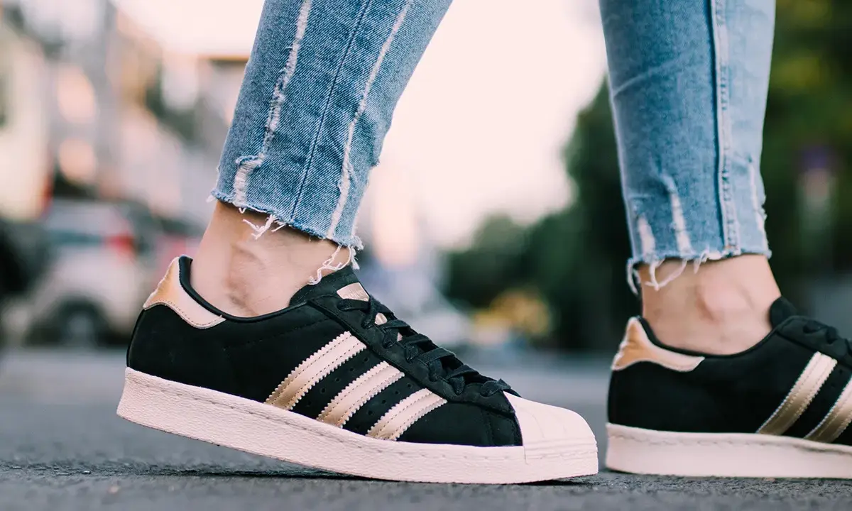 Nogawki jeansów i czarne **modele butów adidas z lat 2000.** ze złotymi paskami i różową podeszwą.