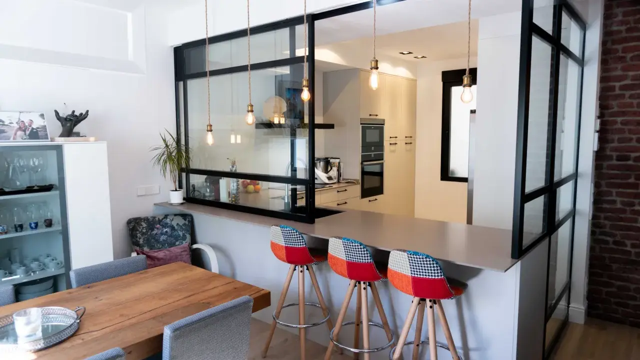 Tabique cocina semiabierta al salón con barra y taburetes coloridos. La cocina moderna se integra al salón a través de un panel de cristal y metal.