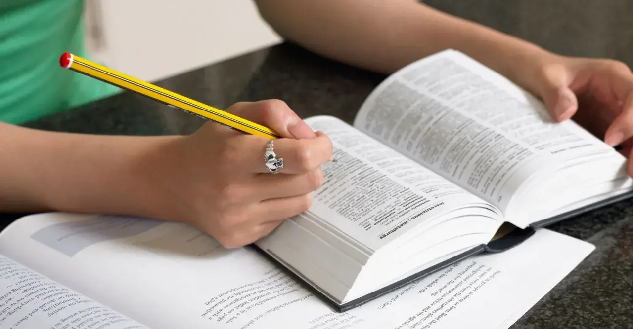 Mani che studiano un dizionario, scrivendo con una matita. Scoprire parole italiane affascinanti è un viaggio.