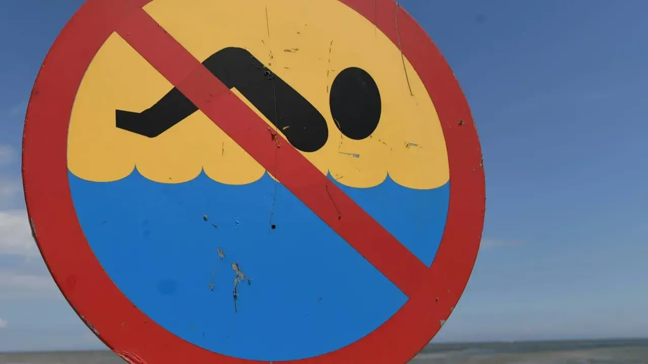 Znak zakazu kąpieli na plaży. Zamknięte kąpieliska nad morzem, mimo pięknej pogody i widocznej w oddali osoby w wodzie.