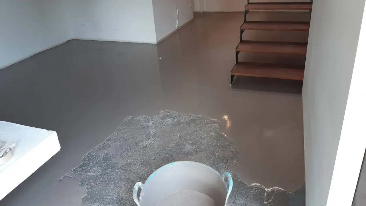 Pavimento continuo de hormigón gris recién aplicado, con una escalera de madera al fondo y un cubo con material.