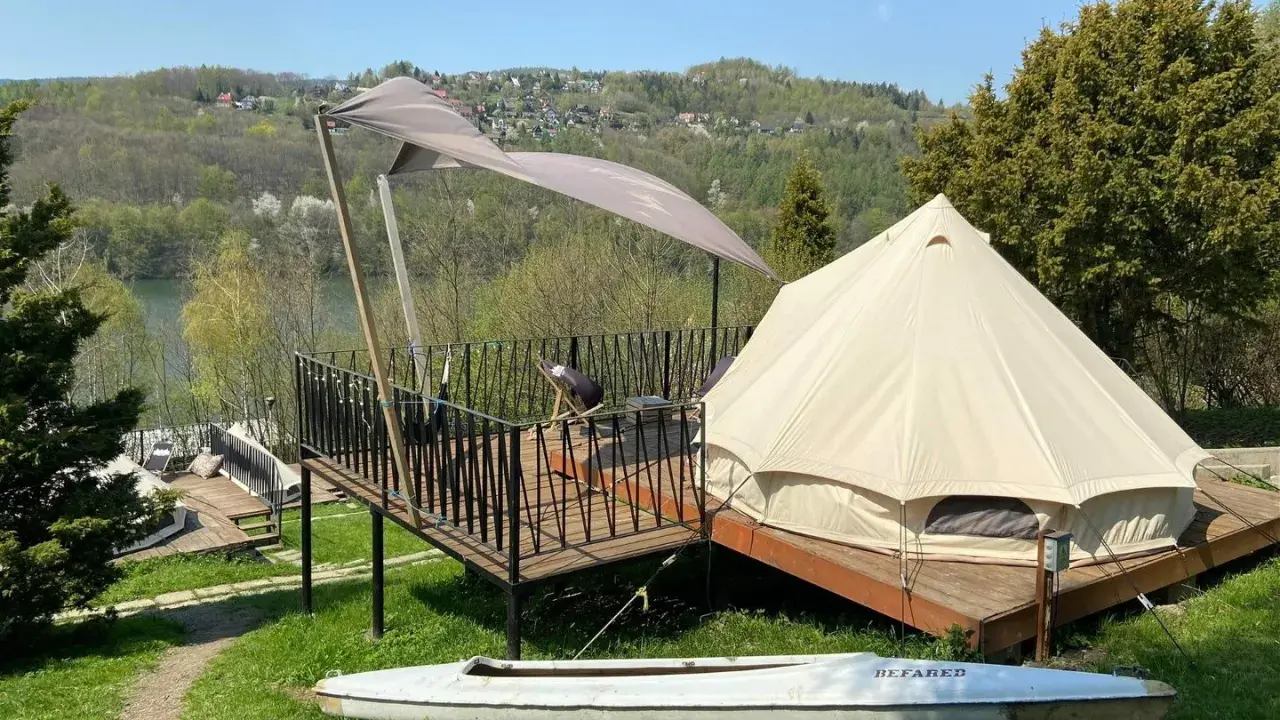 Malowniczy glamping w Polsce z namiotem typu dzwon, tarasem i kajakiem na tle zielonych wzgórz i jeziora.