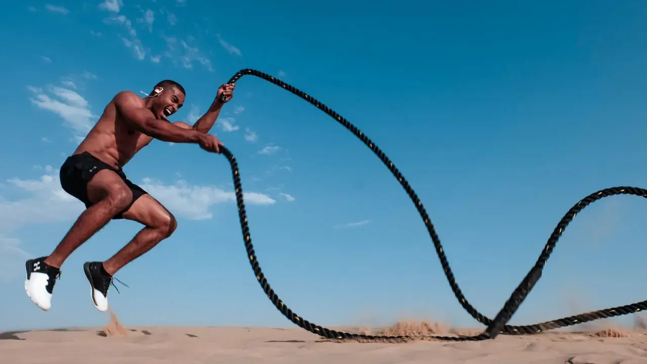 Mann mit Kopfhörern macht ein intensives HIIT-Training mit Seilen im Sand unter blauem Himmel.