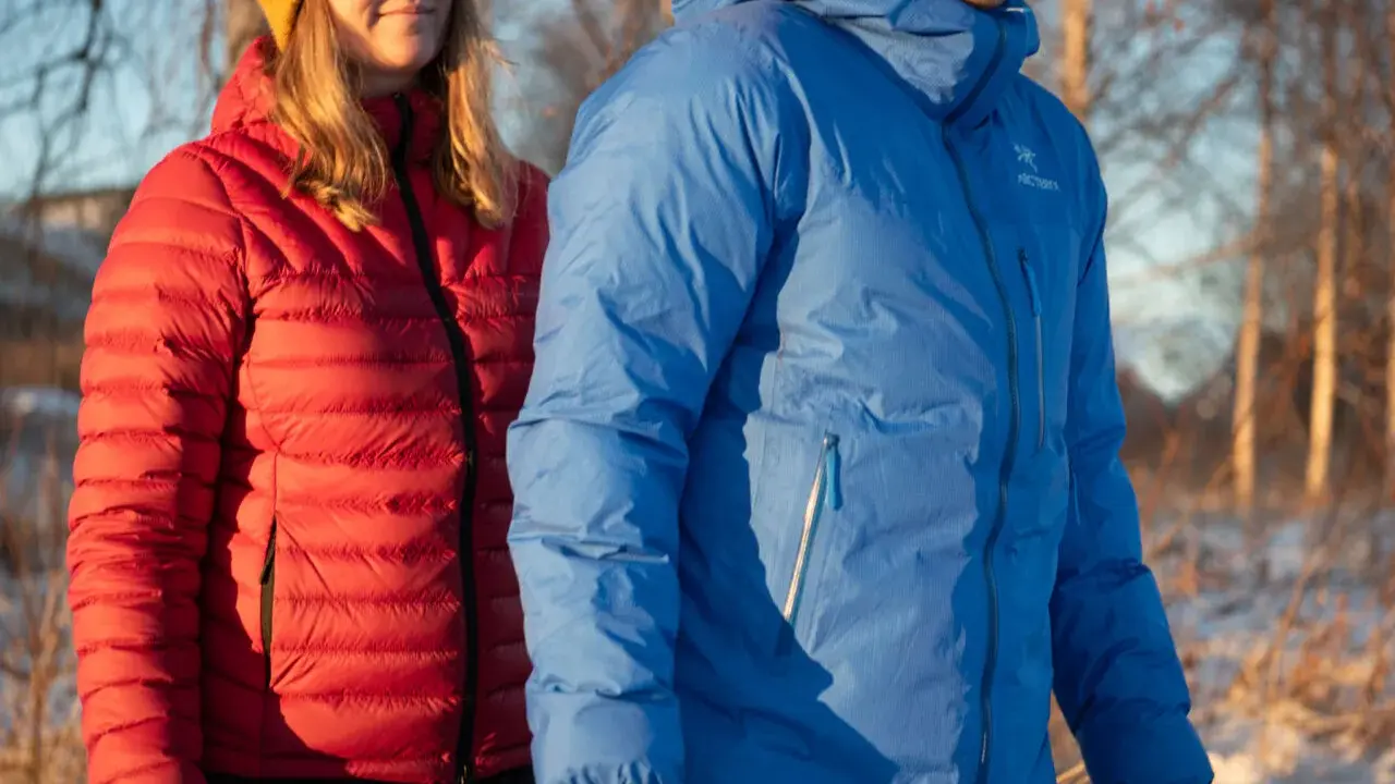 Två personer i vinterkläder, en i röd och en i blå varmaste dunjackan, står utomhus i solljus.