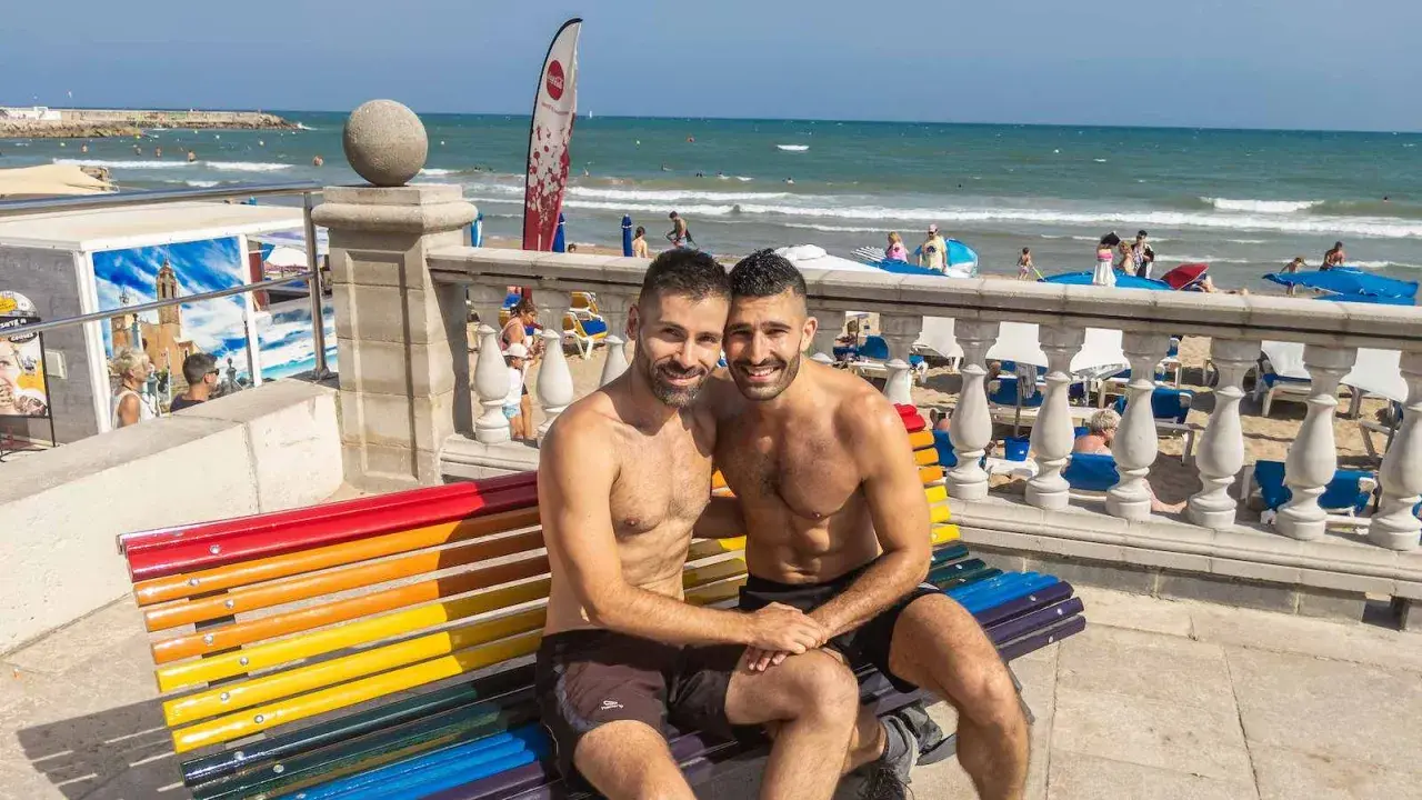 A happy couple enjoys a sunny day at the Sitges gay beach, sitting on a rainbow bench with the ocean in the background.