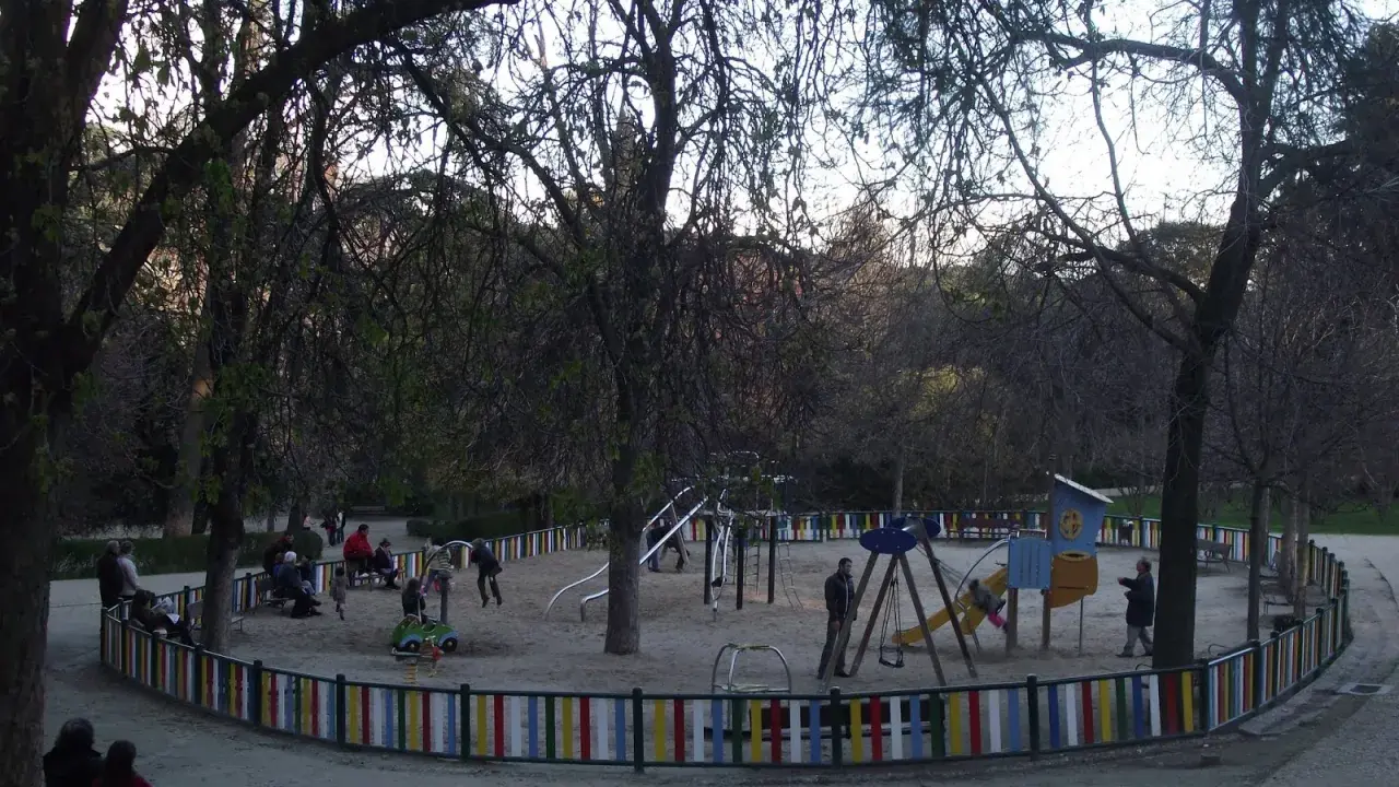 Parque de la Quinta de la Fuente del Berro con niños jugando en columpios y toboganes, rodeado de árboles y una valla colorida.