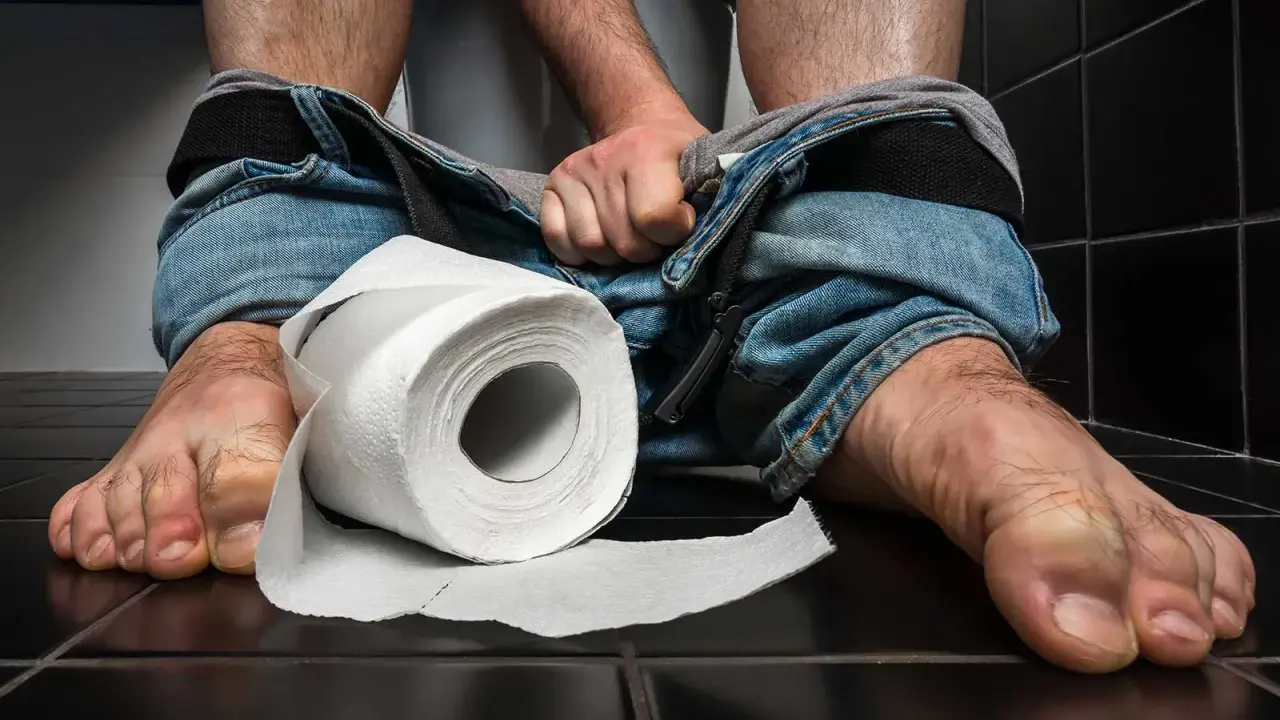 En person sitter på en toalett med neddragna jeans. En rulle toalettpapper ligger bredvid. Tänk på vaccin mot turistdiarré för en tryggare resa.