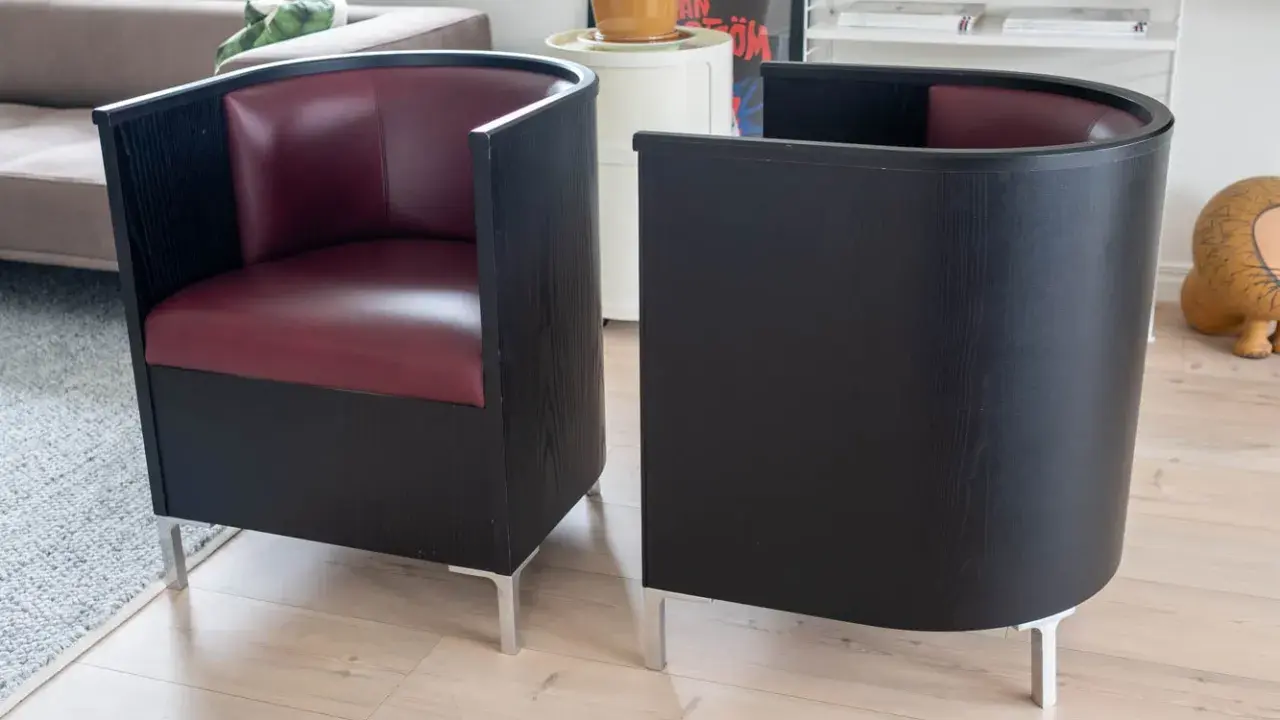Två svarta fåtöljer med vinröda sitsar, designade av Mats Theselius, står på ett ljust trägolv.