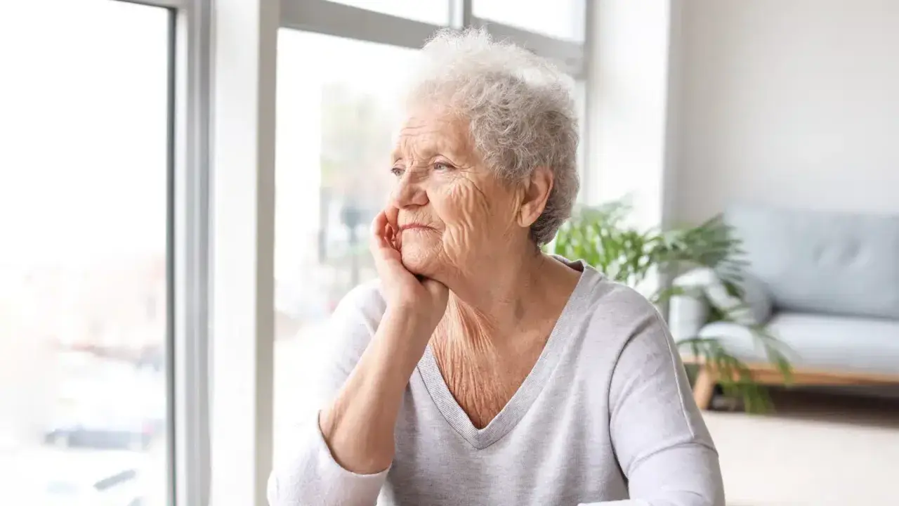 Anziana con mano sul mento, sguardo pensieroso verso la finestra. Potrebbe riflettere sulla pseudodemenza depressiva.