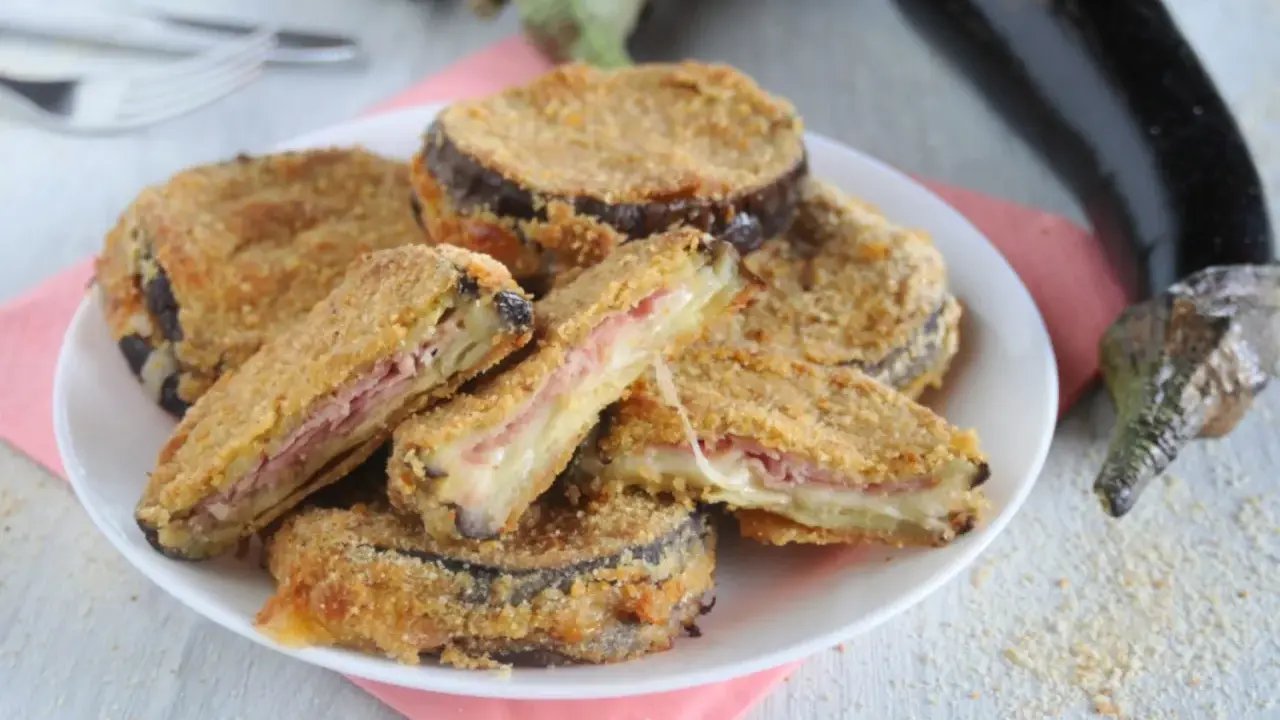 Cordon bleu di melanzane impanati e fritti, ripieni di prosciutto e formaggio filante, serviti su un piatto bianco con melanzane fresche sullo sfondo.