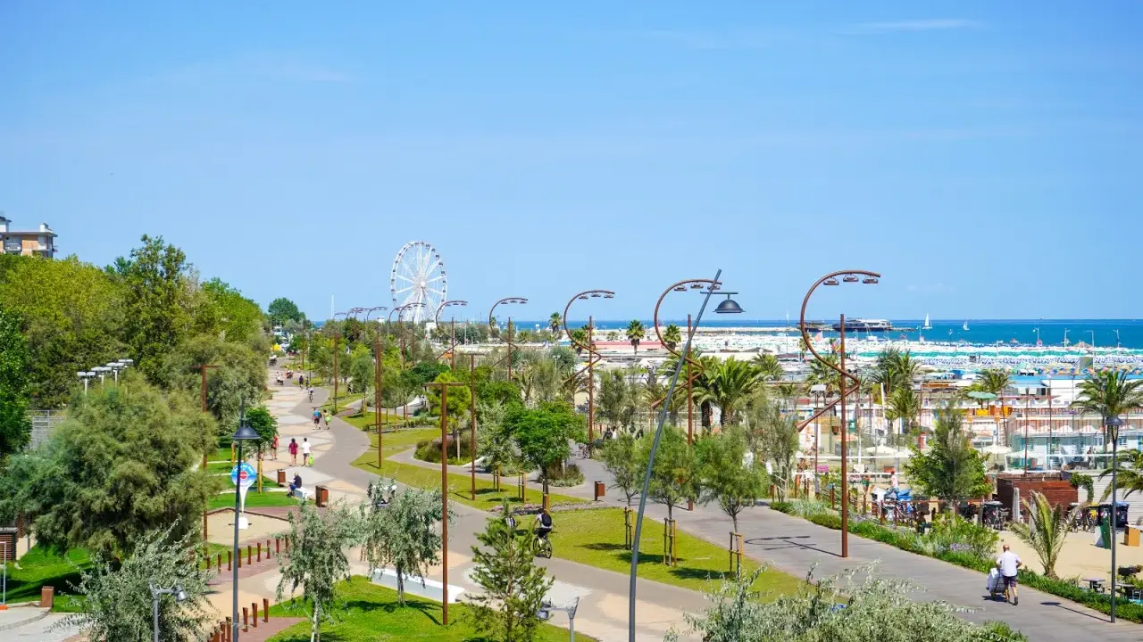Passeggiata nel verde del parco del mare Rimini, con gente che cammina e pedala, e la ruota panoramica sullo sfondo.