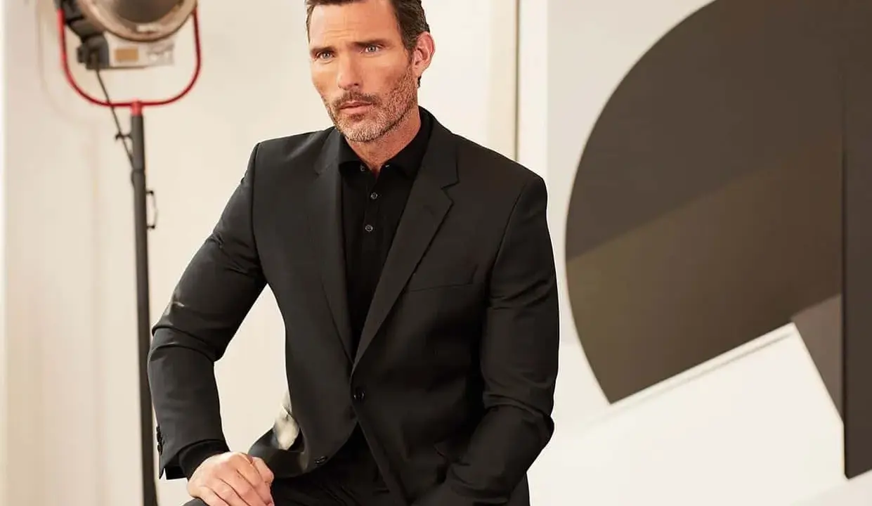 A man in a sharp black suit and polo shirt, a classic funeral outfit for men, poses with a thoughtful expression.